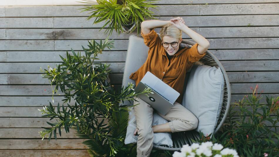 Eine Frau sitzt mit einem Laptop auf dem Schoss auf einer Veranda.