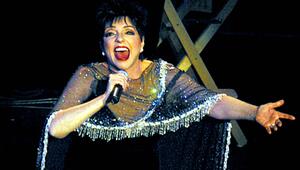 Liza Minnelli singt in ein Mikrofon. Sie trägt eine dunkle Robe und ein Cape mit Glitzerfransen.