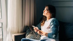 Eine Frau sitzt mit Handy in der Hand auf dem Sofa. Sonne strahlt ihr ins Gesicht.