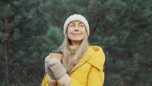 Eine Frau steht in gelber Jacke, mit Mütze und Handschuhen und Kaffeebecher in der Hand im Schnee.