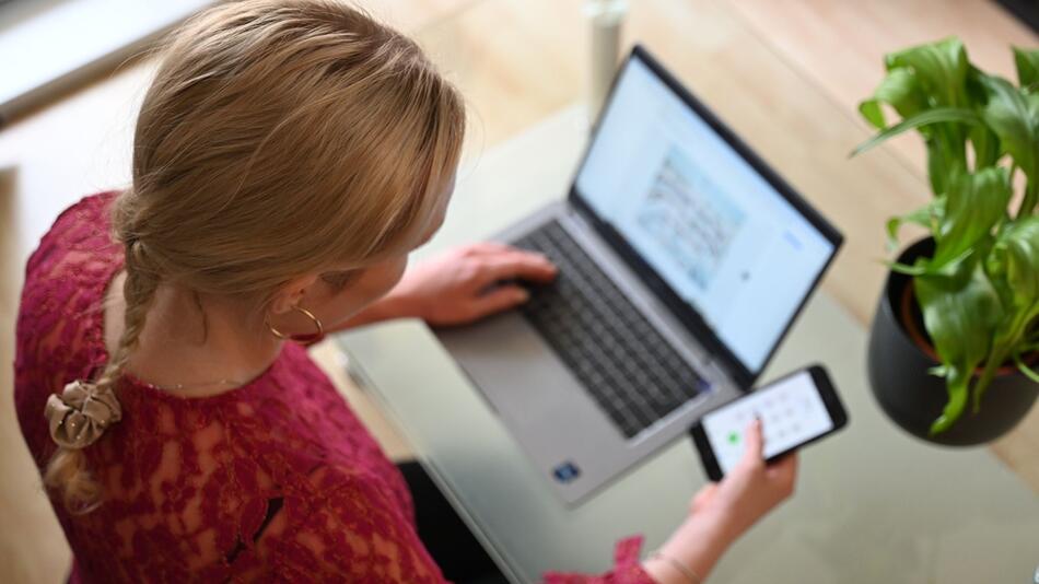 Frau sitzt vor einem Laptop und schaut auf ein Smartphone