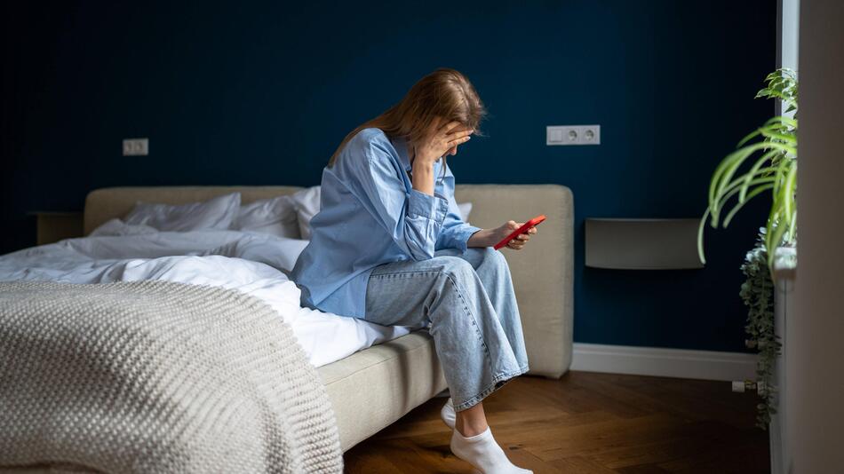 Eine traurige Frau sitzt mit ihrem Smartphone in der Hand auf dem Bett
