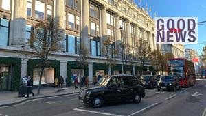 Strassenszene mit einem historischen Gebäude mit Säulen, mehreren Bäumen und Schaufenstern, davor ...