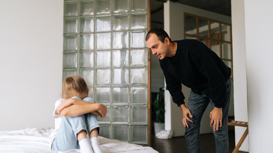 Tochter hat die Arme vor dem Gesicht verschränkt, Vater redet auf sie ein