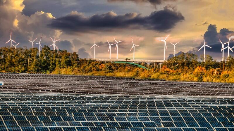 Mehrere Solaranlagen stehen auf einem Feld und im Hintergrund sind Windräder.