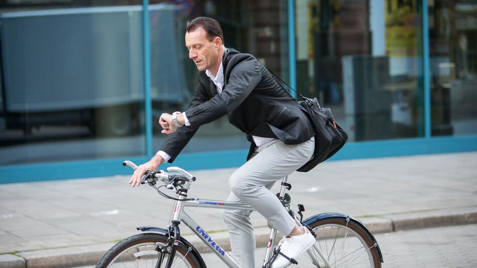 Mann fährt mit Fahrrad durch Stadt und guckt auf Uhr