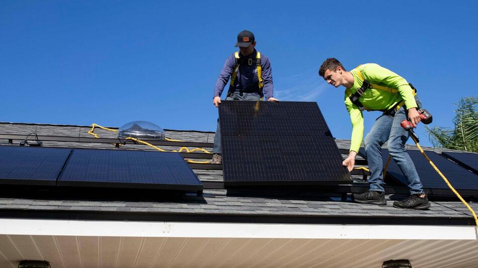 Zwei Männer installieren Solarpaneele auf einem Hausdach. Beide tragen Sicherheitsgurte, einer ...