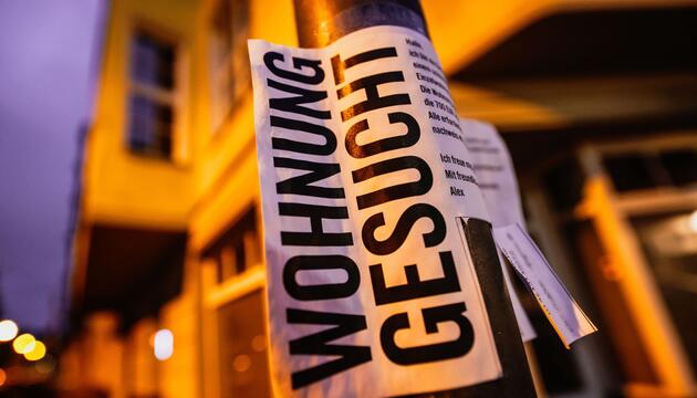 Ein Zettel mit der Aufschrift "Wohnung gesucht" vor Wohnhäusern