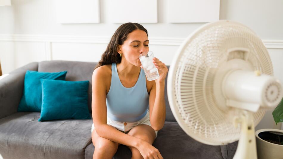 Frau trinkt Eiswasser und lässt sich von Ventilator kühlen