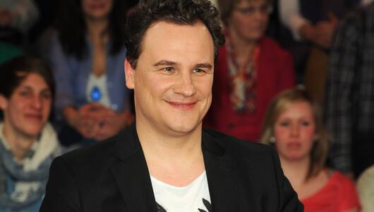 Guido Maria Kretschmer lächelt bei Talkshow. Er trägt einen schwarzem Blazer und ein weisses Shirt.